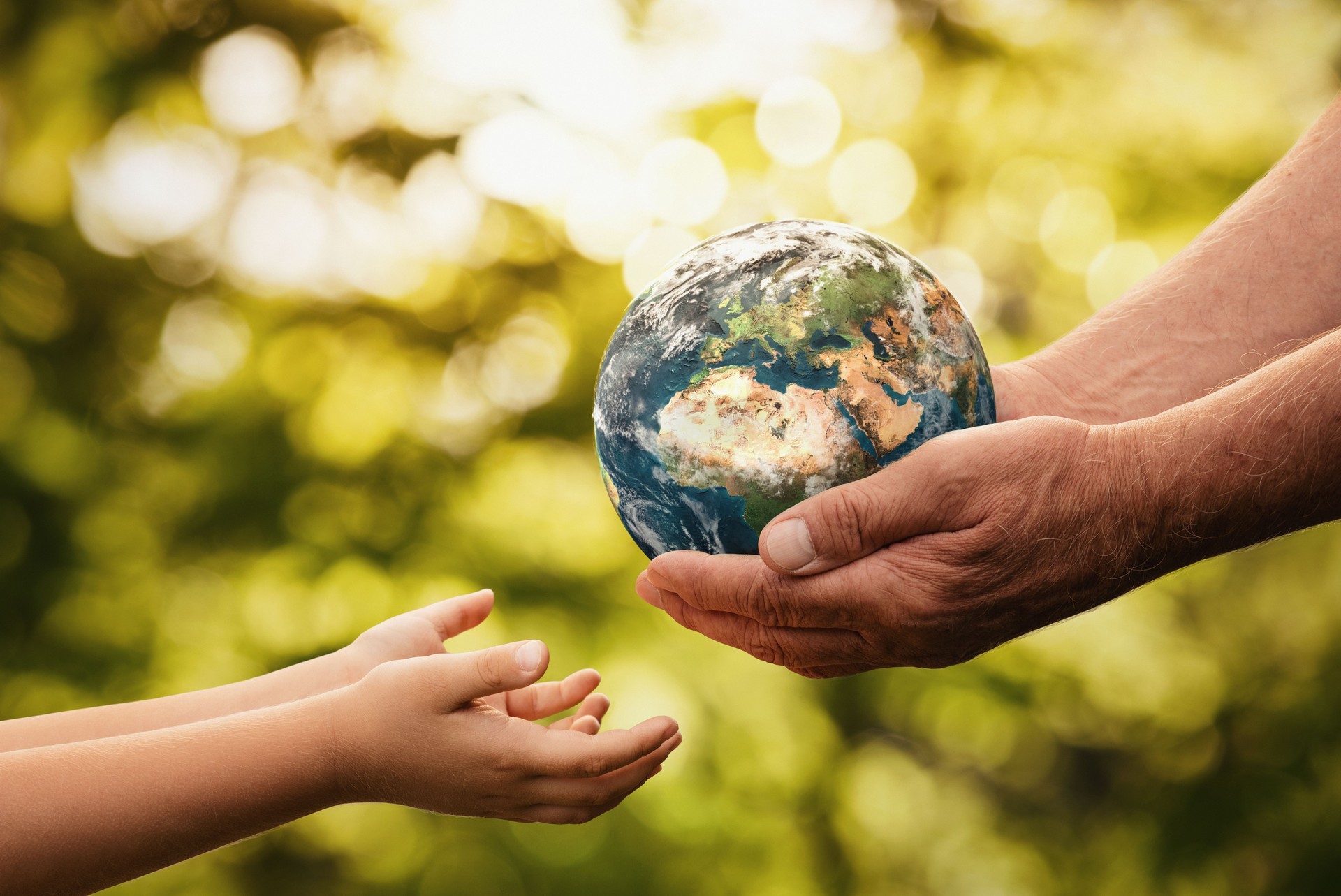 En äldre hand som räcker en jordglob till ett barns utsträckta händer, med en suddig grön bakgrund. Symboliserar överlämning av ansvar för planeten och hållbarhet mellan generationer.